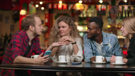 Cheerful-company-of-friends-at-a-bar-drinking-coffee-laughing-and-discussing-while-looking-at-the-smartphone-screen.